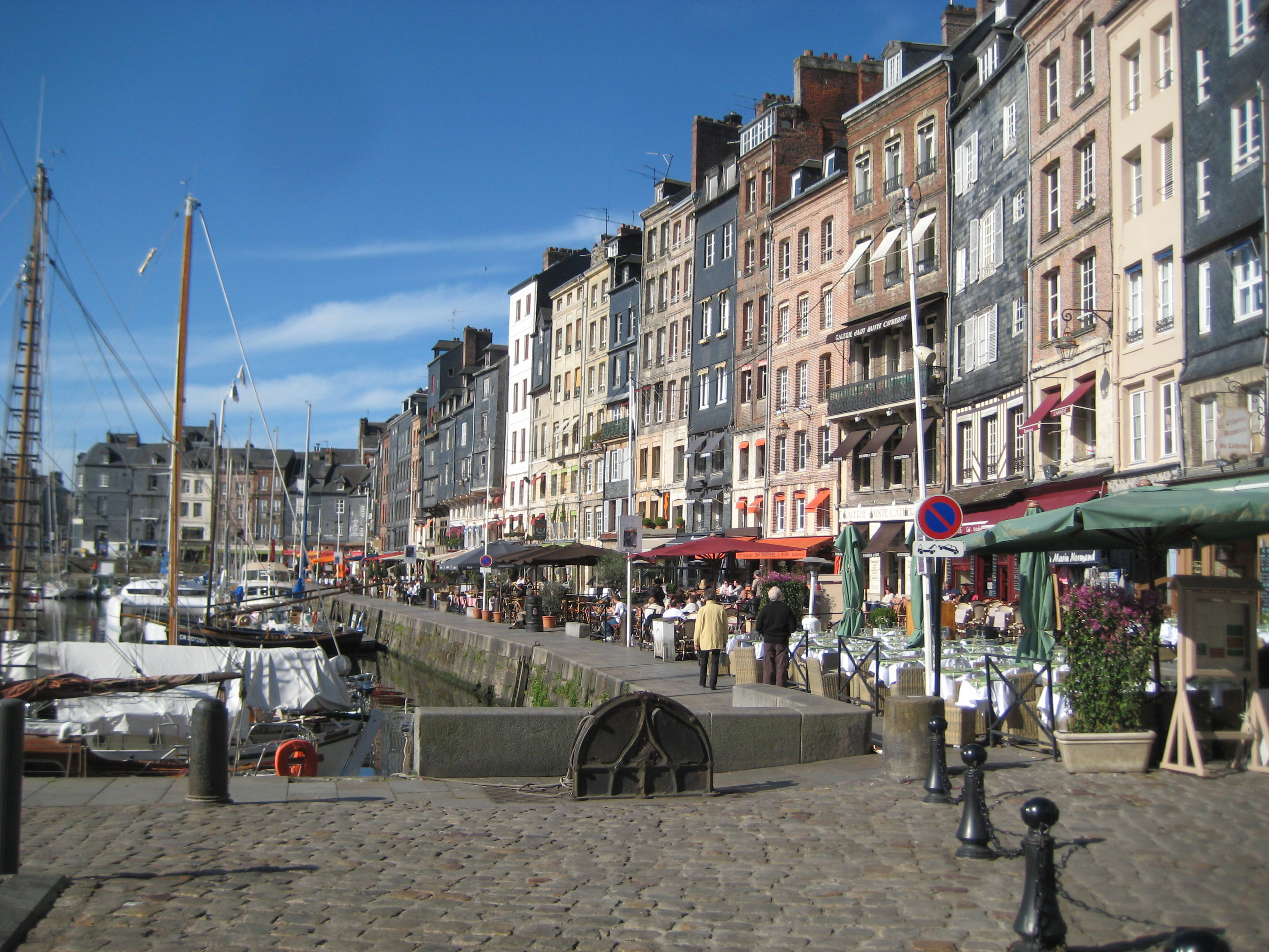 Hamn i Honfleur, Normandie med färgglada historiska hus, uteserveringar och båtar längs kajen under klarblå himmel.