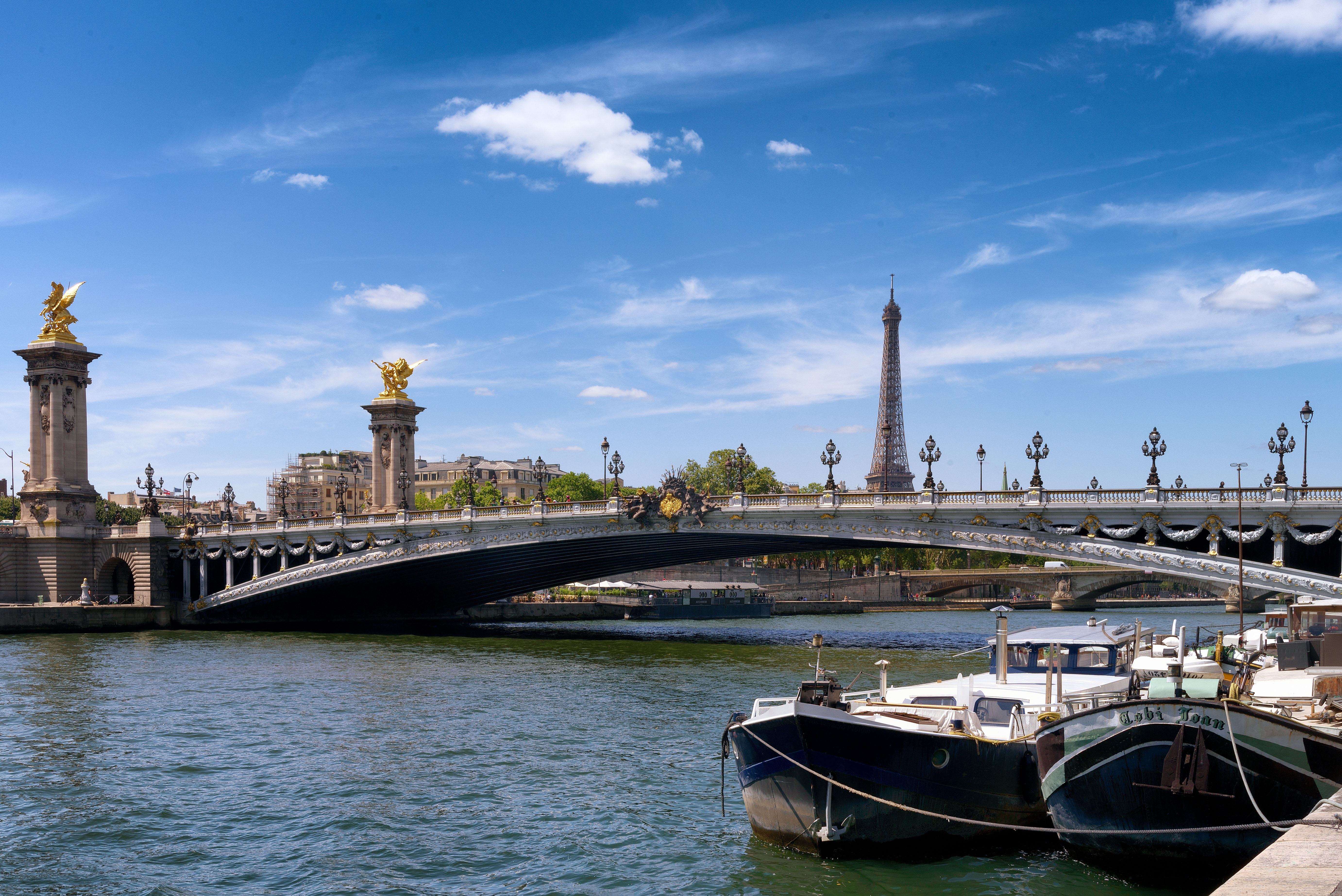 Pont Alexandre III-bron över floden Seine i Paris med Eiffeltornet i bakgrunden och båtar vid kajen under blå himmel.