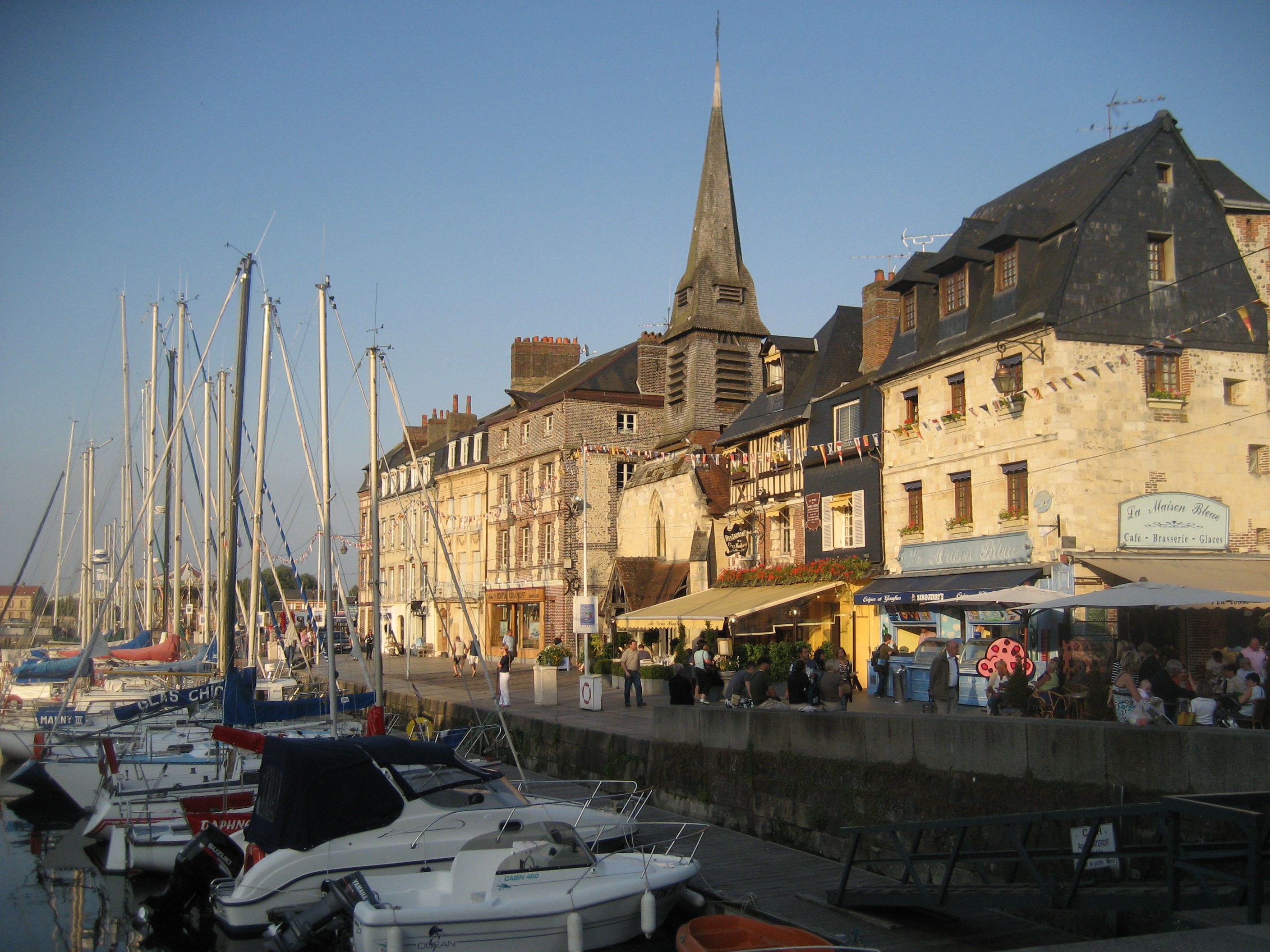 Historiska byggnader och segelbåtar vid hamnen i Honfleur, Normandie, med pittoreska restauranger och kyrkspira under klar himmel.