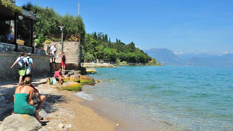 Strandpromenad vid Gardasjön i Italien med klart blått vatten, grönskande omgivning och besökare som njuter av solen.