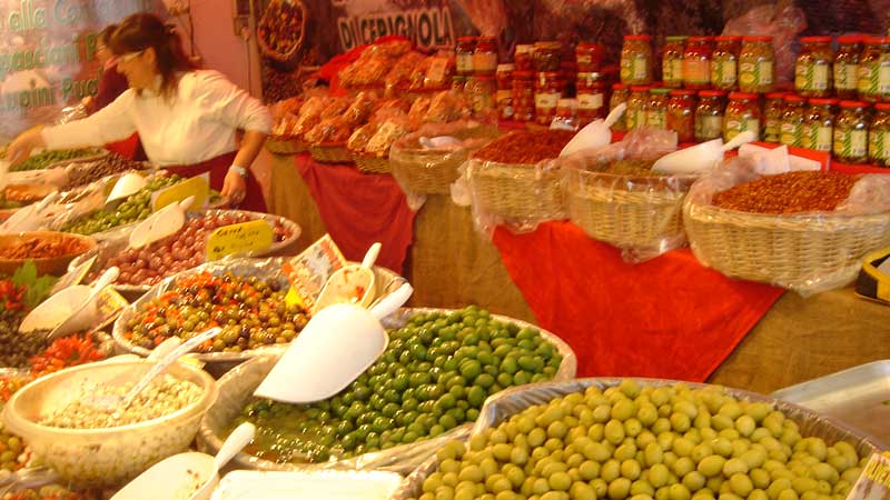 Färgstark marknadsstånd i Italien med korgar fyllda av oliver, torkade tomater och inlagda grönsaker samt burkar med delikatesser.