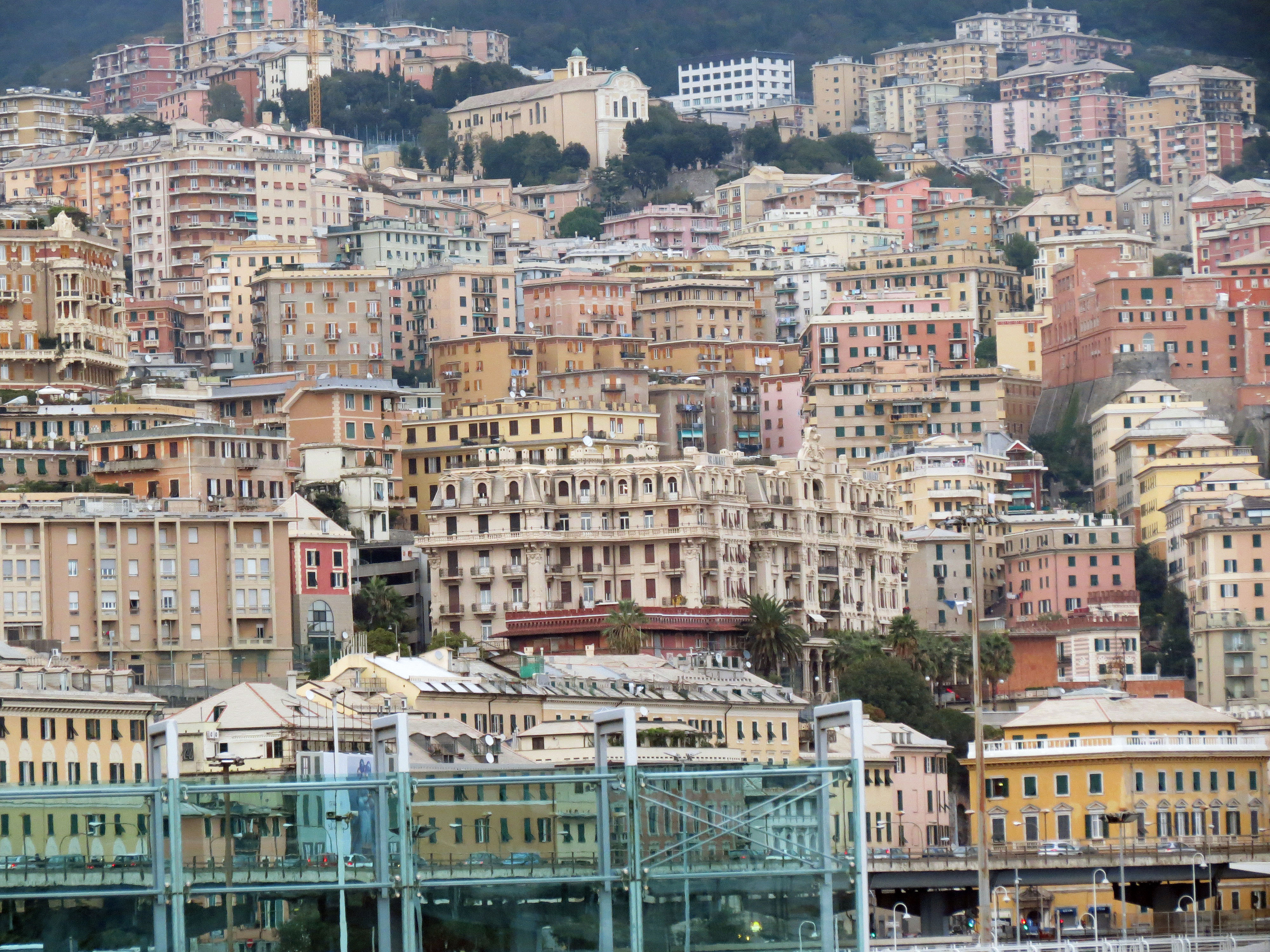 Panoramavy över tätbebyggda historiska och moderna byggnader i Genua, Italien, med kuperad bakgrund och arkitektonisk mångfald.