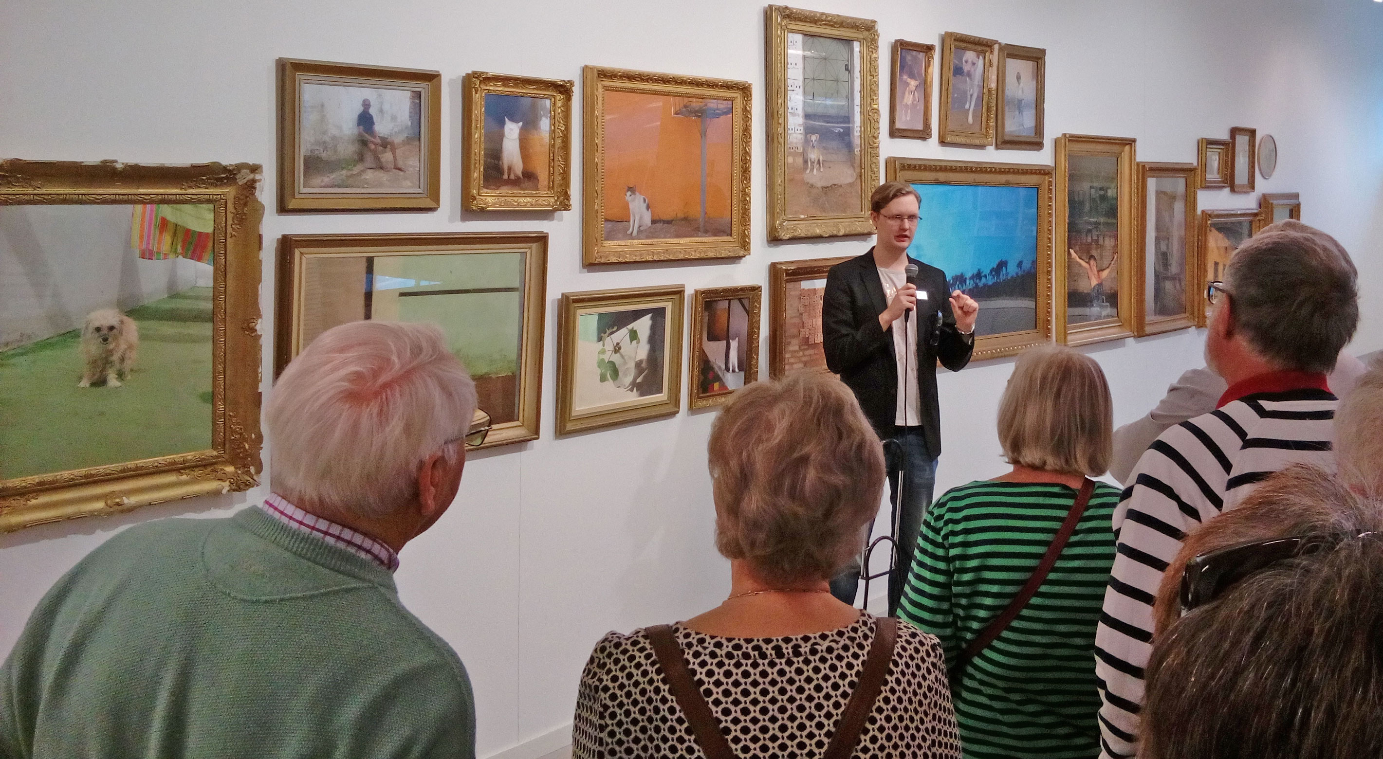 Guidad visning på Lars Lerins Sandgrund i Karlstad. En grupp besökare står framför en vägg med flera inramade målningar i olika storlekar, föreställande landskap, djur och människor.